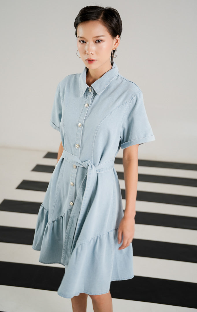 Dress denim wanita lengan pendek warna biru muda (light blue) dengan kerah, kancing depan, sabuk ikat di pinggang, dan detail hem ruffle bergelombang dari This Is April.