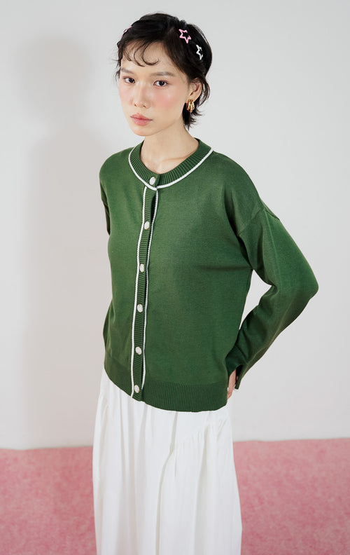 Model mengenakan This Is April Forest Cardigan warna dark green, kardigan rajut lengan panjang dengan detail garis putih kontras (contrast tipping) dan kancing depan.