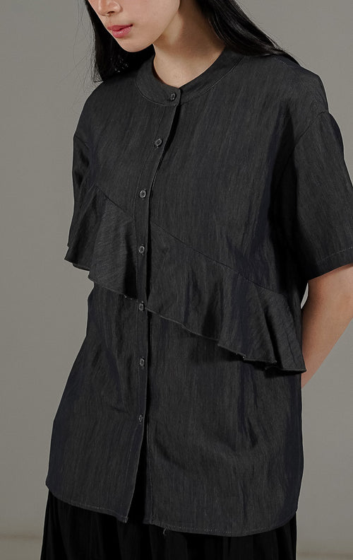 Karina Top Dark Grey dari This Is April, kemeja lengan pendek wanita warna abu abu tua dengan detail kerah bulat dan aksen ruffle asimetris di bagian depan.