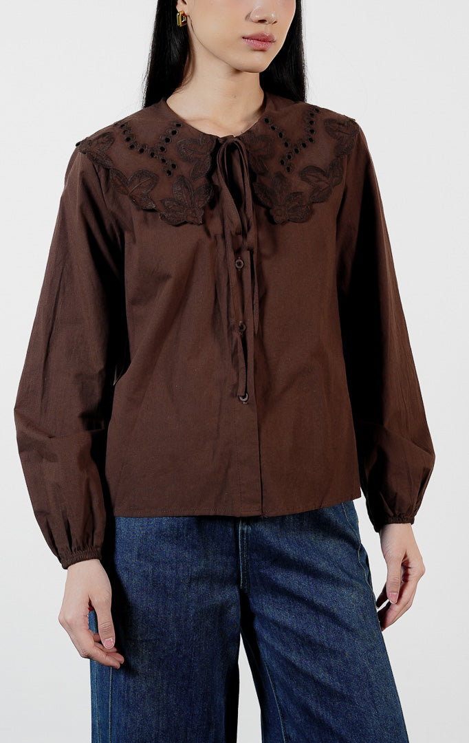 Pakaian wanita model blouse lengan panjang bahan katun dengan detail kerah bordir motif lubang (eyelet broderie) dan tepi gelombang (scalloped edges) warna cokelat tua Lioren Top untuk gaya kasual anggun.