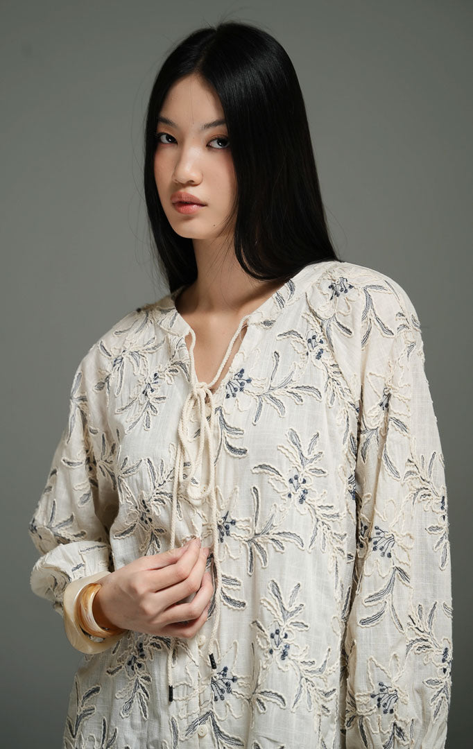 Blus wanita gaya bohemian lengan panjang warna ivory (ivory bohemian floral embroidered top) Robena Top dengan detail bordir motif bunga, bahan katun bertekstur, serta penutup kancing dan tali serut depan yang fleksibel.