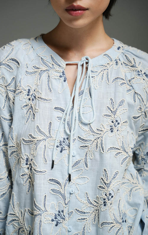 Blus wanita gaya bohemian lengan panjang warna biru muda (light blue bohemian floral embroidered top) Robena Top dengan aksen bordir motif bunga, bahan katun bertekstur, serta kancing dan tali serut depan yang elegan.