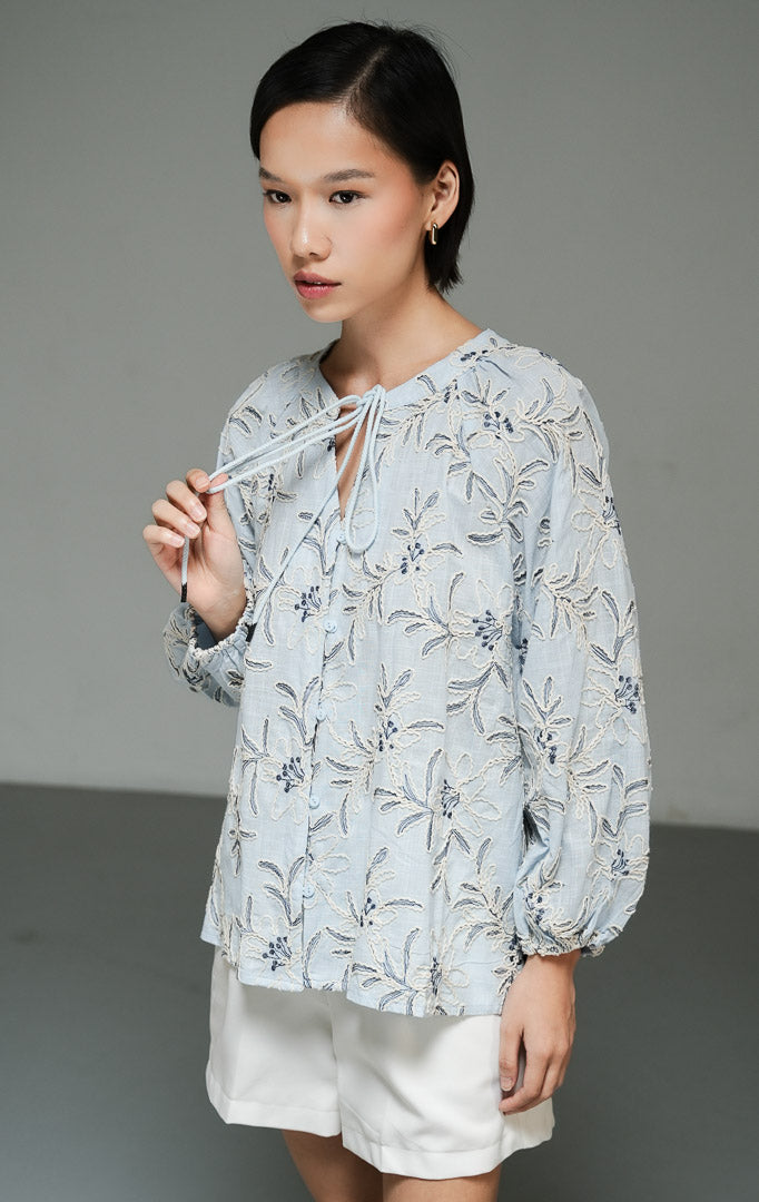 Blus wanita gaya bohemian lengan panjang warna biru muda (light blue bohemian floral embroidered top) Robena Top dengan aksen bordir motif bunga, bahan katun bertekstur, serta kancing dan tali serut depan yang elegan.