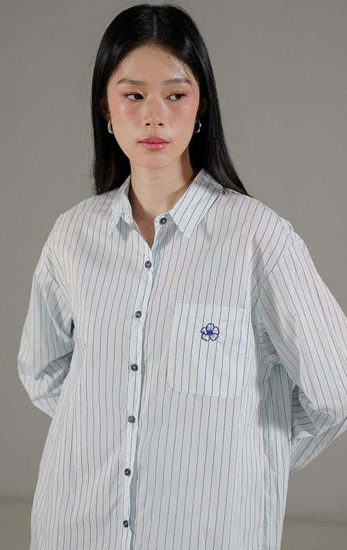 Sion Top Light Blue dari This Is April, kemeja oversize motif salur biru putih dengan saku depan dan detail bordir bunga.