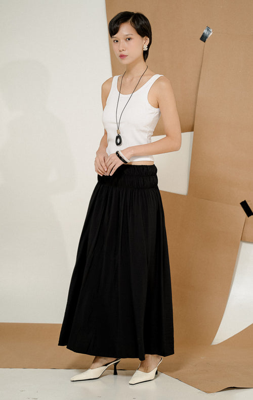 Model berdiri berpose mengenakan Wattson Flowy Ruched Maxi Skirt Black, produk rok panjang wanita model maxi warna hitam dengan detail pinggang elastis berkerut dipadukan tank top putih.