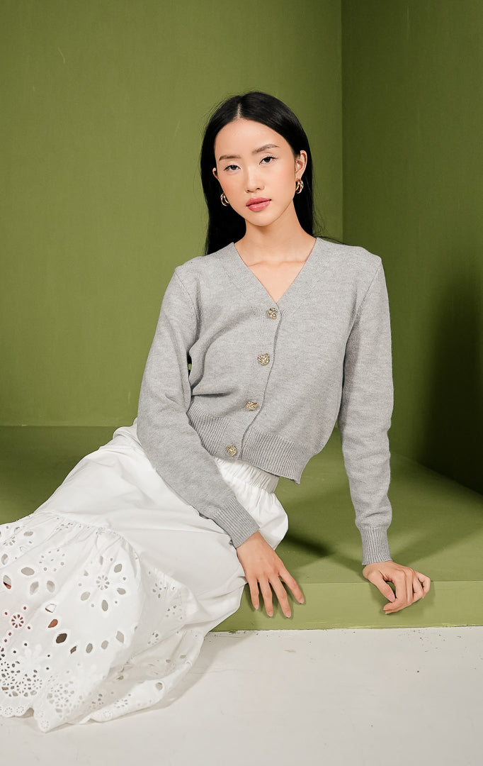 Kardigan rajut wanita Yonna Cardigan model crop warna abu-abu polos lengan panjang, dilengkapi kerah V-neck, detail rib pada ujung lengan dan bagian bawah, serta empat kancing metal berwarna emas di bagian tengah depan.