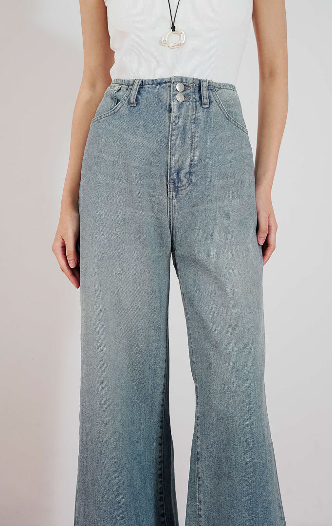 Model wanita mengenakan Zeline High Waist Denim Pants warna biru muda berpotongan lebar dengan detail dua kancing depan, dipadukan dengan tanktop putih.