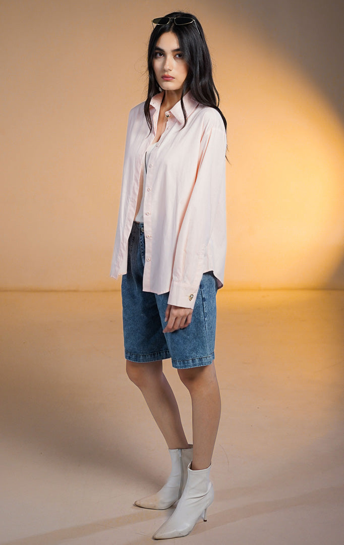 Model wanita memakai Zorrel Top Peach, kemeja lengan panjang warna pink pastel dengan kancing statement emas di kerah, dipadukan dengan celana pendek denim.
