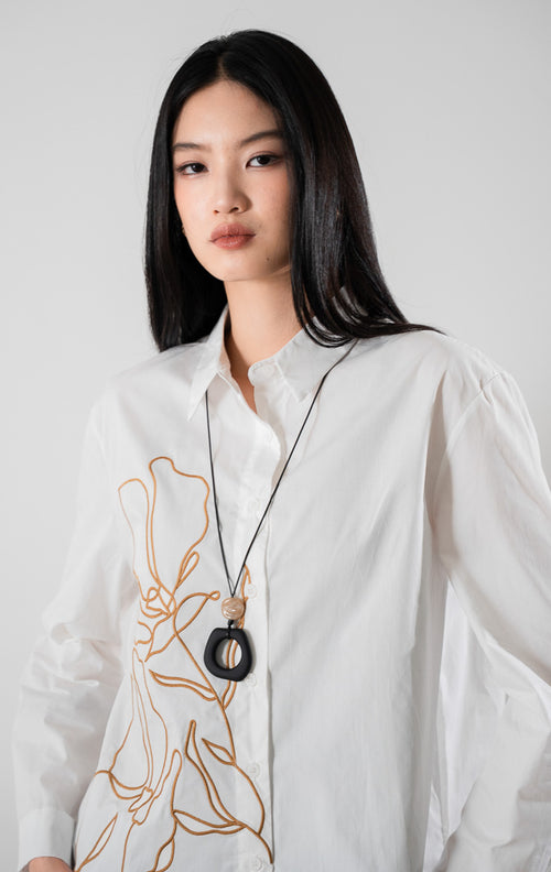 kemeja lengan panjang berwarna putih Adela Top White berbahan katun dengan detail bordir bunga estetis di bagian depan, dipadukan dengan celana cokelat.