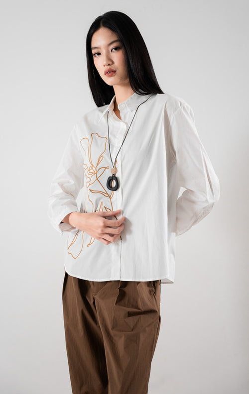 kemeja lengan panjang berwarna putih Adela Top White berbahan katun dengan detail bordir bunga estetis di bagian depan, dipadukan dengan celana cokelat.