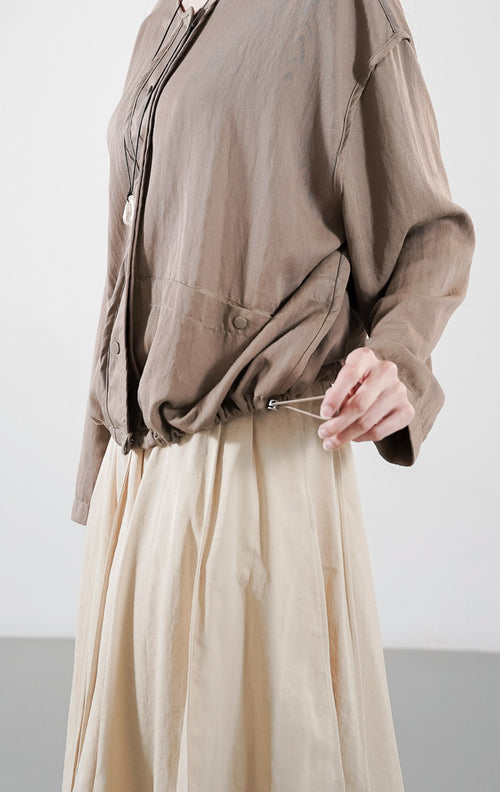 Alexine Jacket kasual warna taupe, jaket ringan dengan detail kancing depan dan tali serut (drawstring), dipadukan dengan rok plisket.