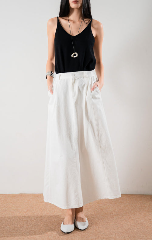 Tampilan bawah rok panjang Almira Skirt White dengan detail lipit depan, kancing, dan saku, dipadukan dengan atasan hitam dan sepatu flat putih.