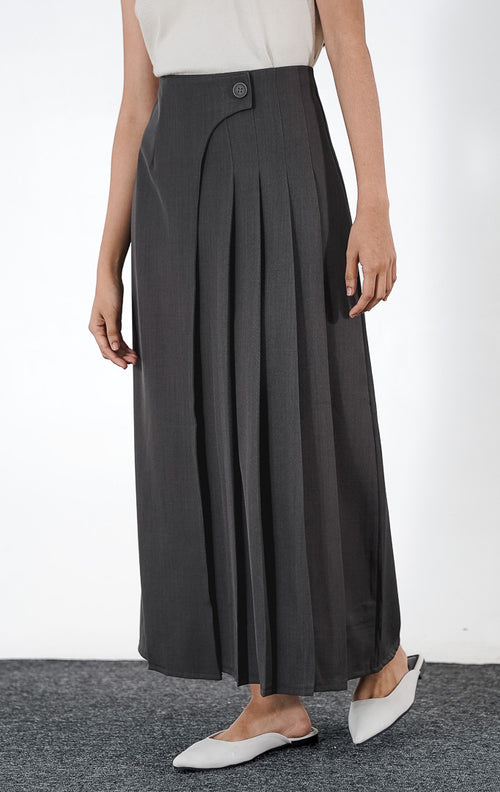 Azalia Skirt warna dark grey, rok panjang plisket berbahan poliester dengan aksen kancing asimetris di pinggang, dipadukan dengan sepatu putih.