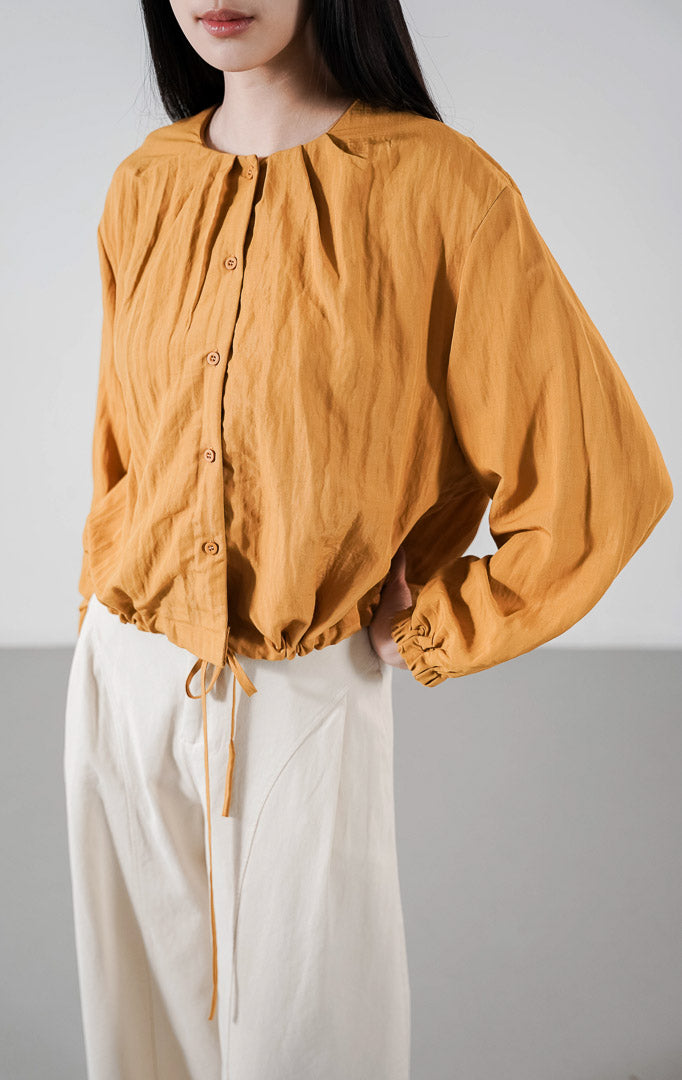 Callum Top warna mustard, atasan lengan panjang berkerah bulat dengan kancing depan dan detail tali serut (drawstring) di pinggang, dipadukan dengan celana putih tulang.