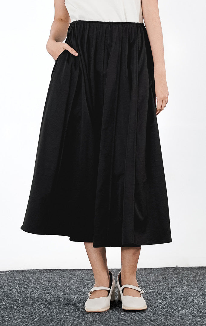 Catarina Skirt warna hitam, rok midi potongan A-line dengan detail lipit (gathered) dan saku samping, dipadukan dengan sepatu Mary Jane putih.