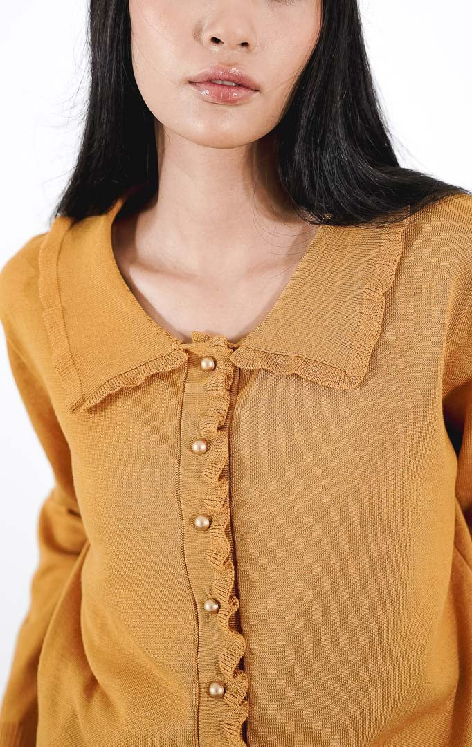  Catriona Top warna mustard, atasan rajut lengan panjang dengan detail kerah ruffle dan kancing emas bulat, dipadukan dengan celana kulot warna krem.