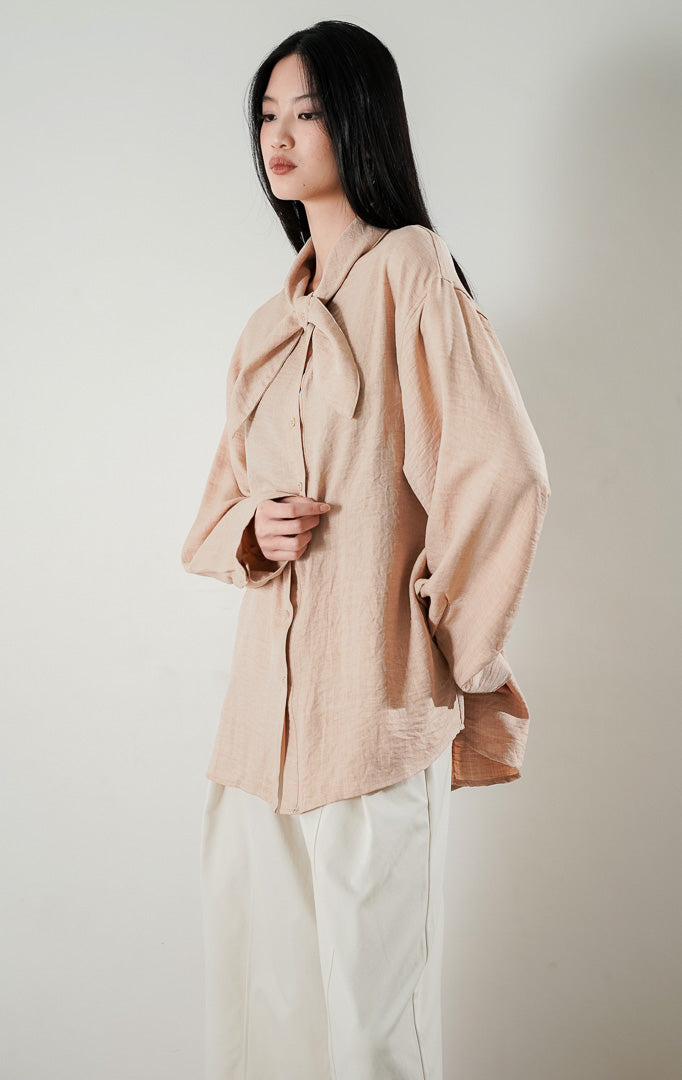 kemeja lengan panjang berwarna khaki atau krem Cristina Top berbahan katun dengan detail kerah pita di bagian leher, dipadukan dengan celana kulot cream dan sepatu heels silver