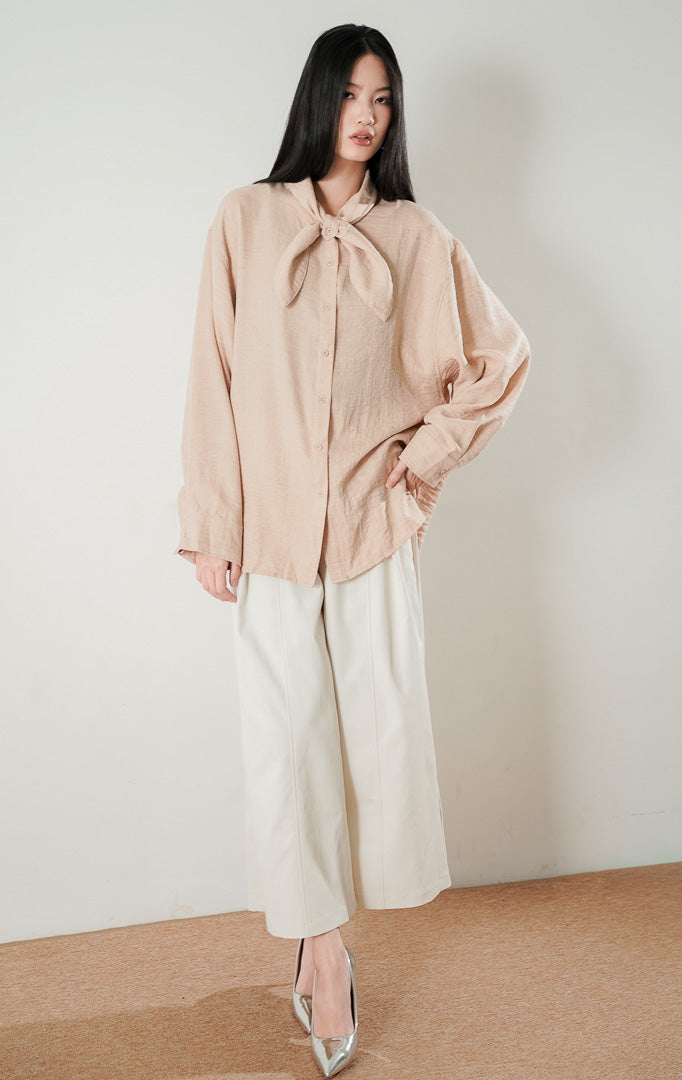 kemeja lengan panjang berwarna khaki atau krem Cristina Top berbahan katun dengan detail kerah pita di bagian leher, dipadukan dengan celana kulot cream dan sepatu heels silver
