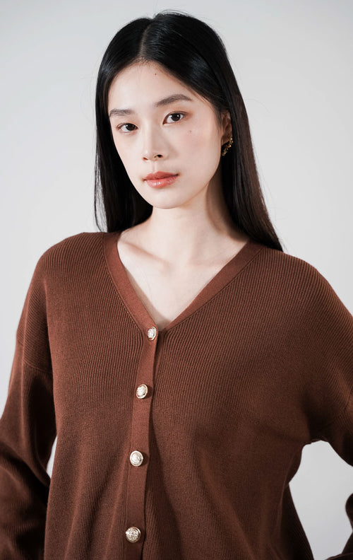  Delphine Cardigan warna dark brown, kardigan rajut V-neck lengan panjang dengan kancing emas bulat, dipadukan dengan rok plisket krem.