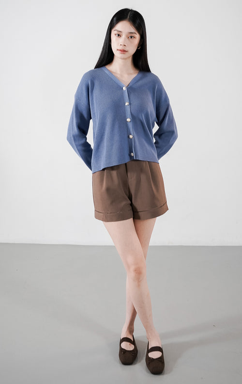 Delphine Cardigan warna dusty blue, kardigan rajut V-neck lengan panjang dengan kancing minimalis, dipadukan dengan celana pendek cokelat.
