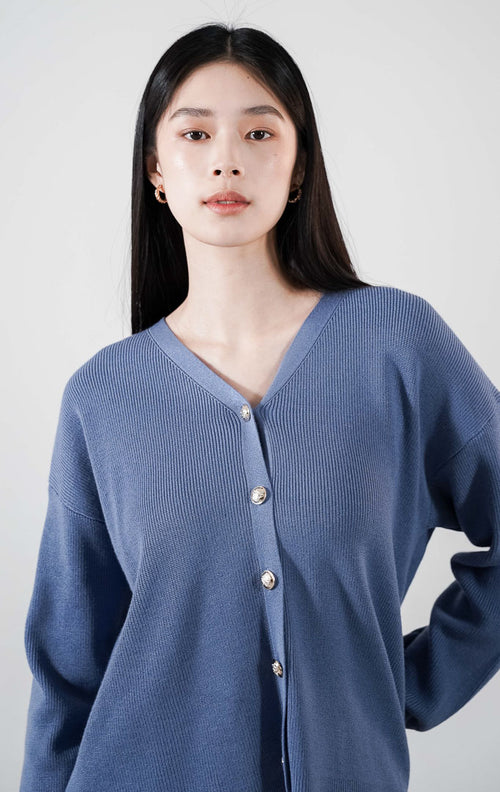 Delphine Cardigan warna dusty blue, kardigan rajut V-neck lengan panjang dengan kancing minimalis, dipadukan dengan celana pendek cokelat.
