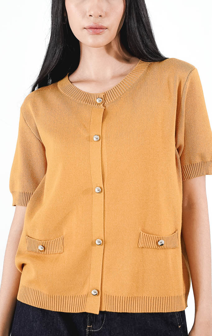 Eliea Top warna mustard, kardigan rajut lengan pendek berkerah bulat dengan detail kancing perak dan dua saku dekoratif di depan.