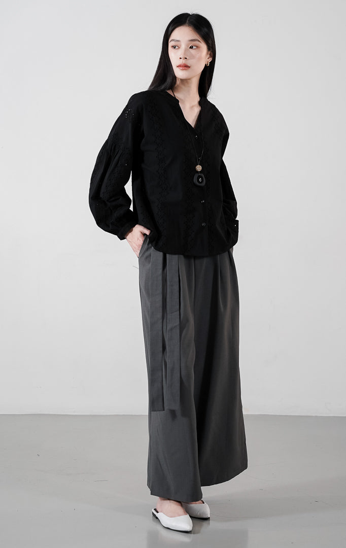 Marcelo Top warna black, atasan katun lengan balon dengan detail bordir eyelet vertikal dan kerah V-neck, dipadukan dengan celana abu-abu.