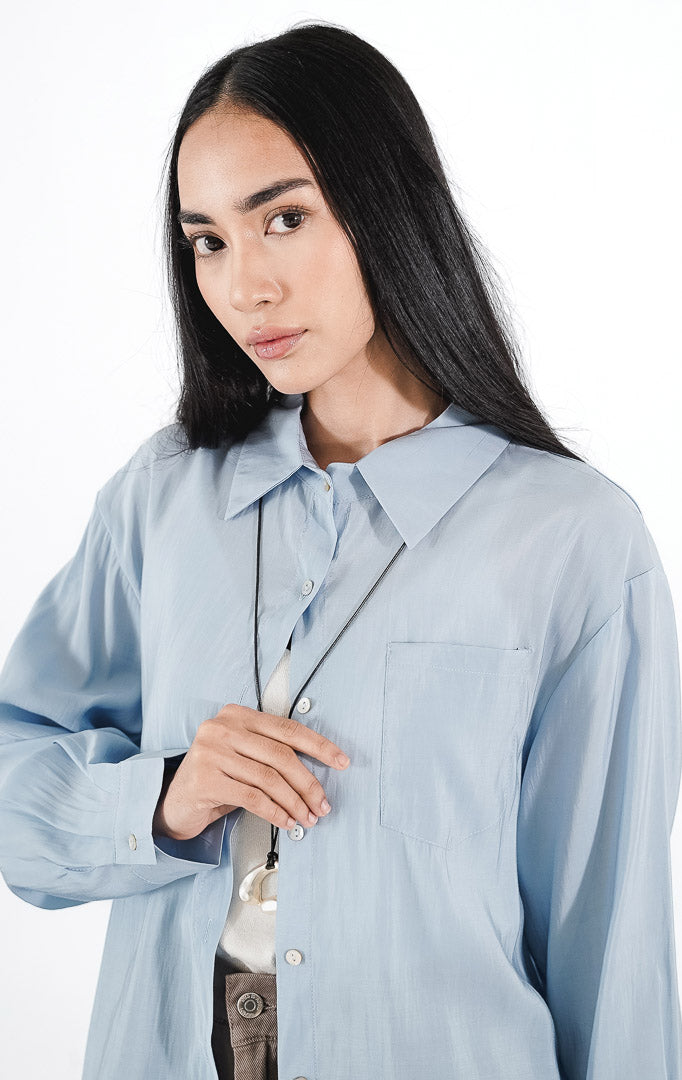 Matea Top warna sky blue, kemeja katun lengan panjang siluet oversized dengan saku dada kiri dan kancing putih minimalis.