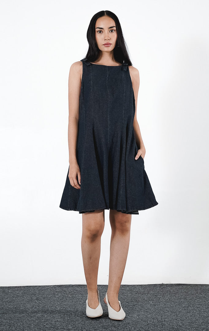  Minuite Dress warna indigo, gaun mini denim tanpa lengan dengan siluet A-line, detail pita di bahu, dan saku samping fungsional.1