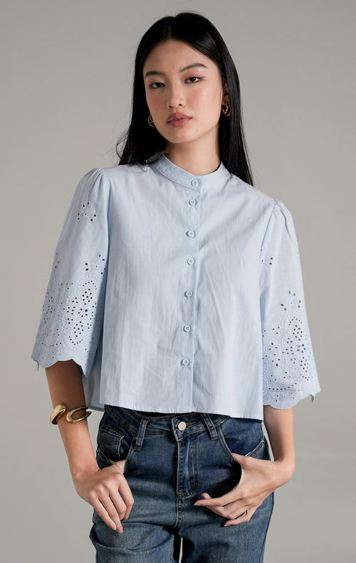  Rochelle Top Light Blue, kemeja katun biru muda dengan detail bordir eyelet pada lengan pendek yang melebar, dipadukan dengan celana jeans wide-leg.