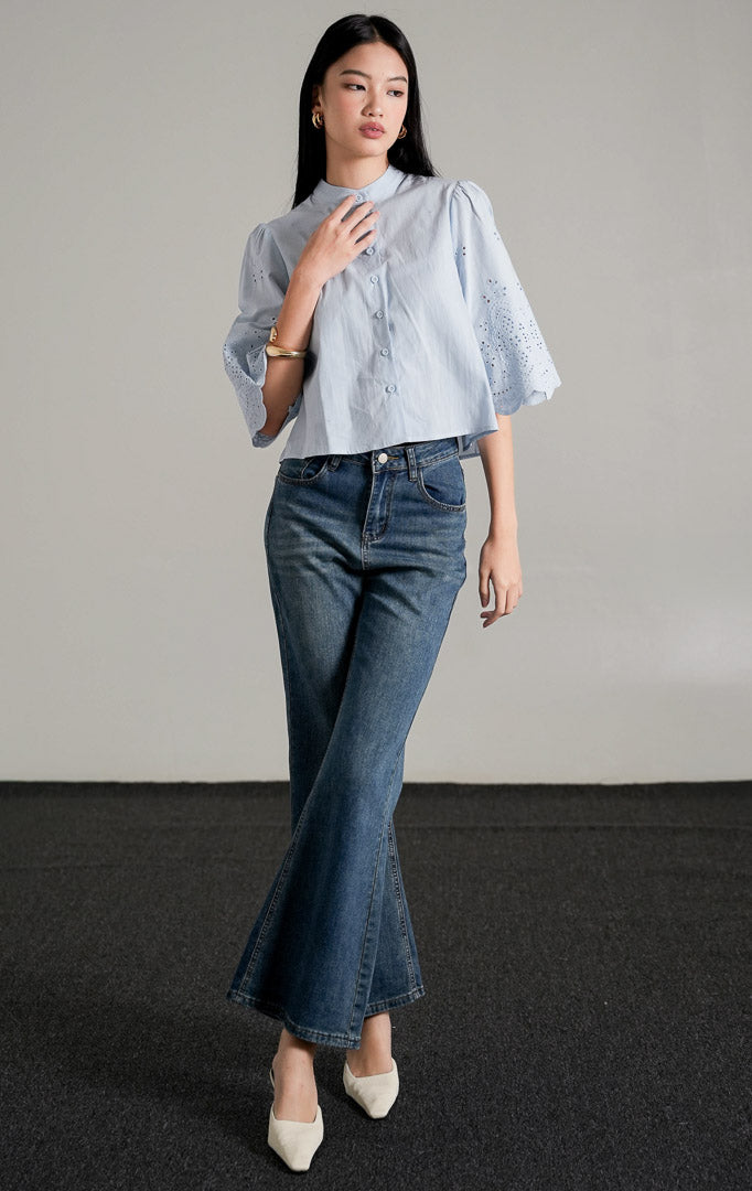  Rochelle Top Light Blue, kemeja katun biru muda dengan detail bordir eyelet pada lengan pendek yang melebar, dipadukan dengan celana jeans wide-leg.