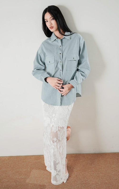 Close up wajah dan tubuh bagian atas model wanita yang mengenakan Adriana Top, kemeja light denim bermotif garis dengan kancing depan dan dua saku dada fungsional.