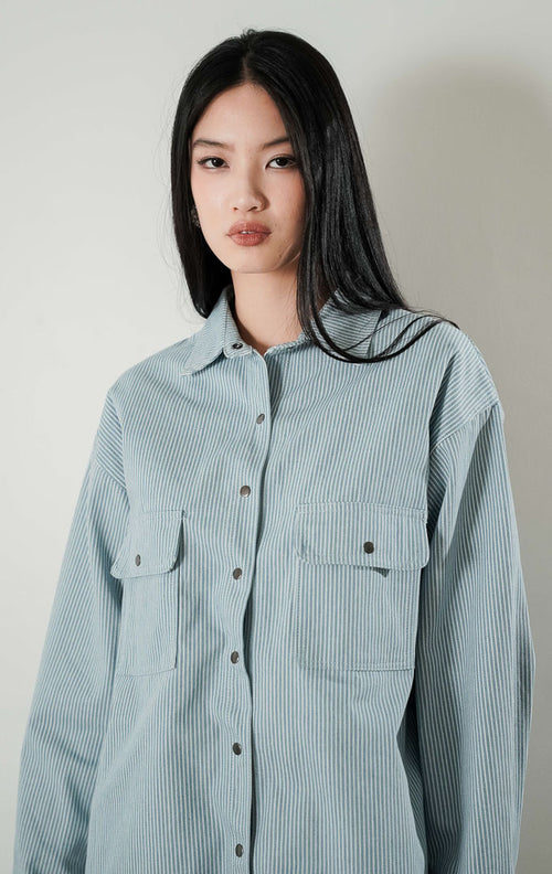 Close up wajah dan tubuh bagian atas model wanita yang mengenakan Adriana Top, kemeja light denim bermotif garis dengan kancing depan dan dua saku dada fungsional.