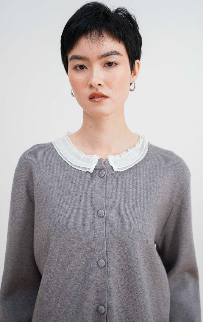 Model mengenakan Aiko Cardigan warna grey, menampilkan detail kerah ruffle lace putih yang cantik, material rajut halus, dan kancing bungkus di bagian depan.