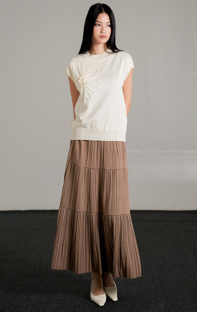 Ailynn Sleeveless Knit Top warna ivory yang memiliki detail applique bunga besar di bagian dada, dipadukan dengan rok panjang beraksen lipit warna cokelat.