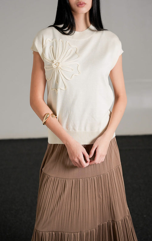 Ailynn Sleeveless Knit Top warna ivory yang memiliki detail applique bunga besar di bagian dada, dipadukan dengan rok panjang beraksen lipit warna cokelat.