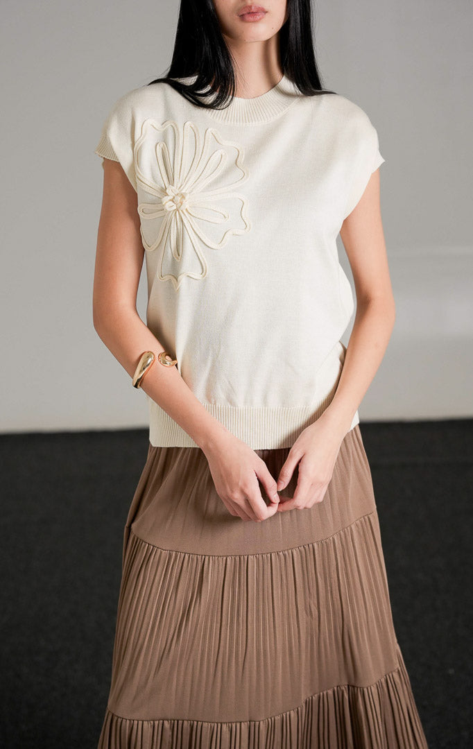 Ailynn Sleeveless Knit Top warna ivory yang memiliki detail applique bunga besar di bagian dada, dipadukan dengan rok panjang beraksen lipit warna cokelat.