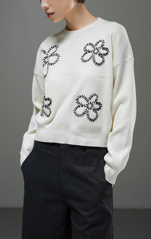 Sweater rajut wanita Amora Sweater warna ivory krem muda lengan panjang dengan motif bordir bunga besar warna hitam, kerah bulat, dan detail ribbed hem. 