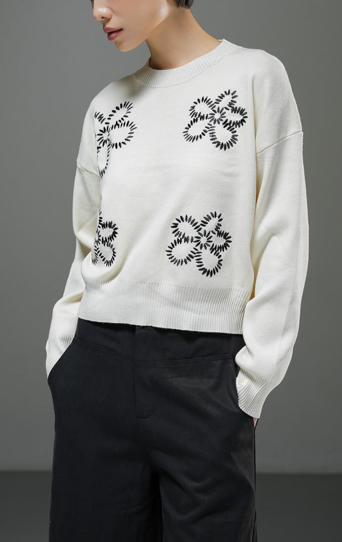 Sweater rajut wanita Amora Sweater warna ivory krem muda lengan panjang dengan motif bordir bunga besar warna hitam, kerah bulat, dan detail ribbed hem. 