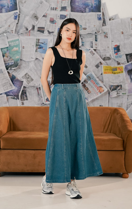 Model wanita mengenakan This Is April Anaya Skirt Denim, rok jeans panjang model maxi potongan A-line warna biru medium wash. Rok memiliki saku depan fungsional, detail jahitan vertikal, dan dipadukan dengan atasan hitam serta sepatu sneakers untuk gaya casual.