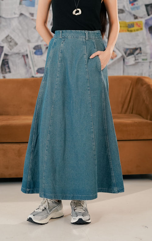 Model wanita mengenakan This Is April Anaya Skirt Denim, rok jeans panjang model maxi potongan A-line warna biru medium wash. Rok memiliki saku depan fungsional, detail jahitan vertikal, dan dipadukan dengan atasan hitam serta sepatu sneakers untuk gaya casual.