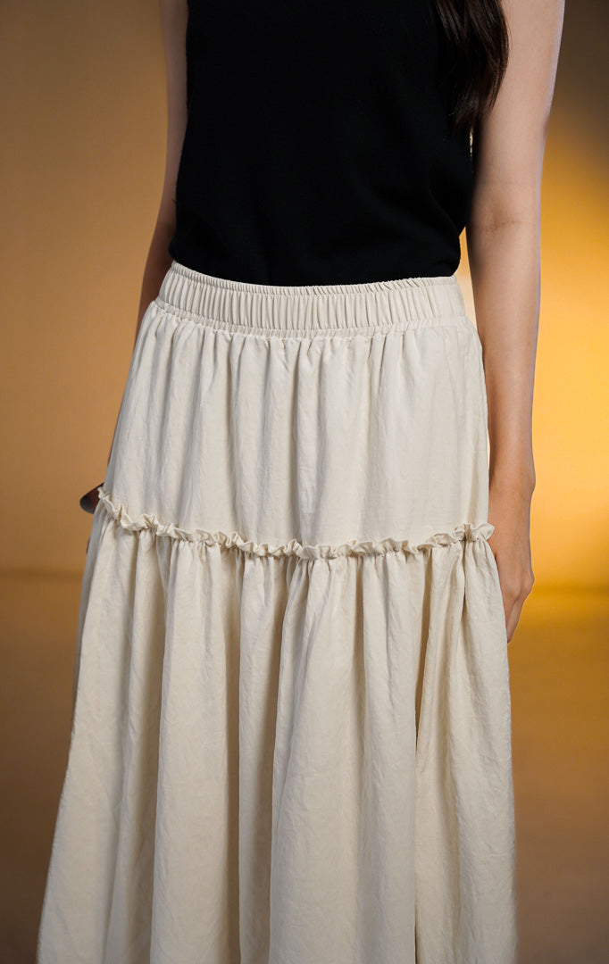 Model wanita mengenakan rok midi susun (tiered skirt) warna ivory bahan peach skin yang flowy dengan atasan tank top hitam
