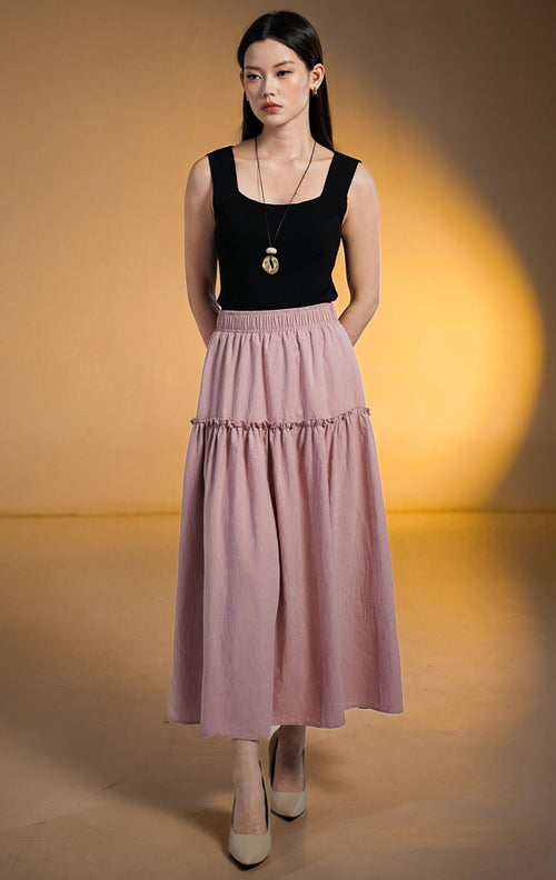 Model wanita mengenakan rok midi susun (tiered skirt) warna merah muda (pink) bahan peach skin yang lembut, dipadukan dengan atasan hitam dan aksesori gelang emas.