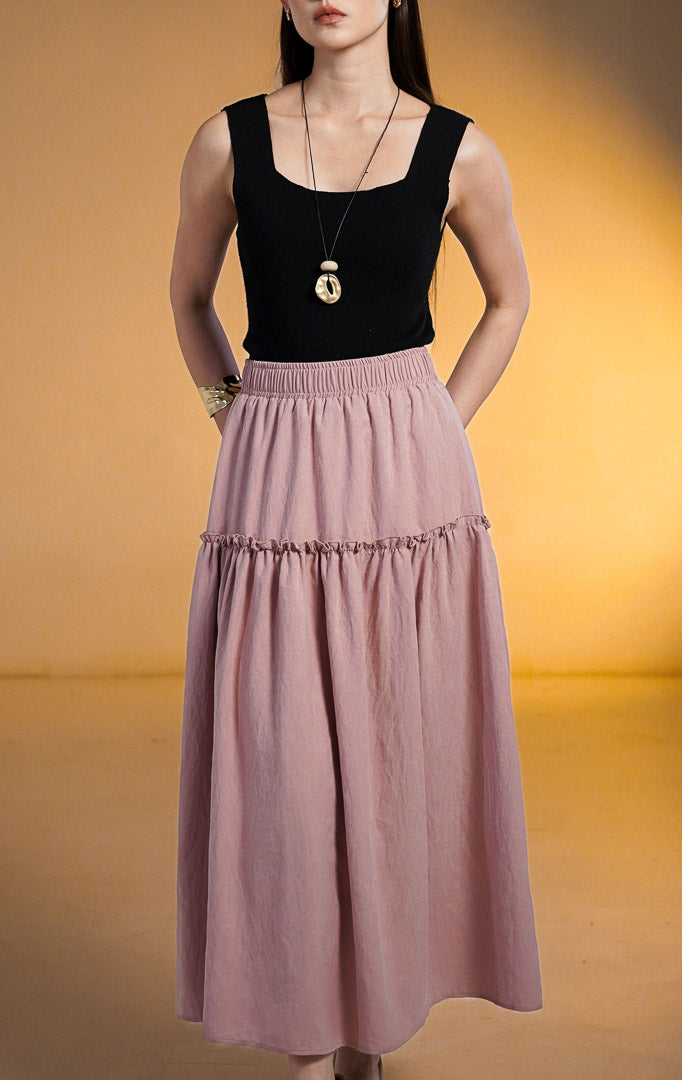 Model wanita mengenakan rok midi susun (tiered skirt) warna merah muda (pink) bahan peach skin yang lembut, dipadukan dengan atasan hitam dan aksesori gelang emas.