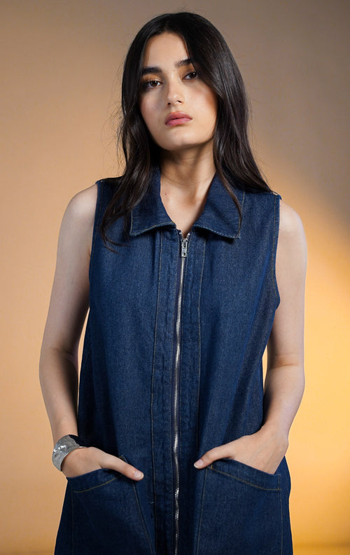 Model wanita mengenakan mini dress denim gelap tanpa lengan dengan resleting depan (zip up) dan dua saku depan besar, dipadukan dengan sepatu boots tinggi hitam.