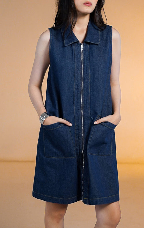 Model wanita mengenakan mini dress denim gelap tanpa lengan dengan resleting depan (zip up) dan dua saku depan besar, dipadukan dengan sepatu boots tinggi hitam.