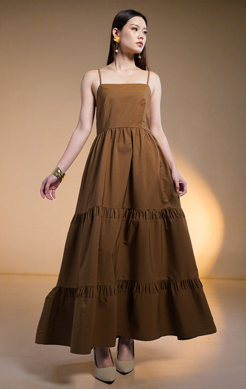 Model wanita mengenakan maxi dress susun (tiered dress) warna cokelat tua (dark brown) dengan tali spageti dan saku samping, dipadukan dengan anting bunga emas.