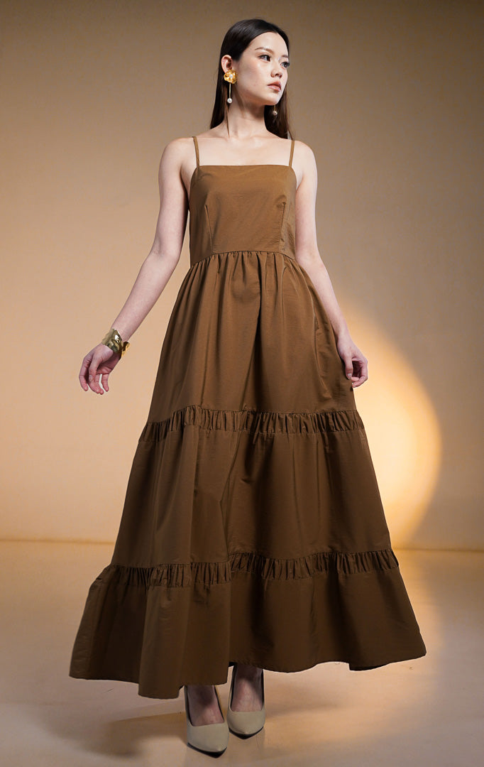 Model wanita mengenakan maxi dress susun (tiered dress) warna cokelat tua (dark brown) dengan tali spageti dan saku samping, dipadukan dengan anting bunga emas.