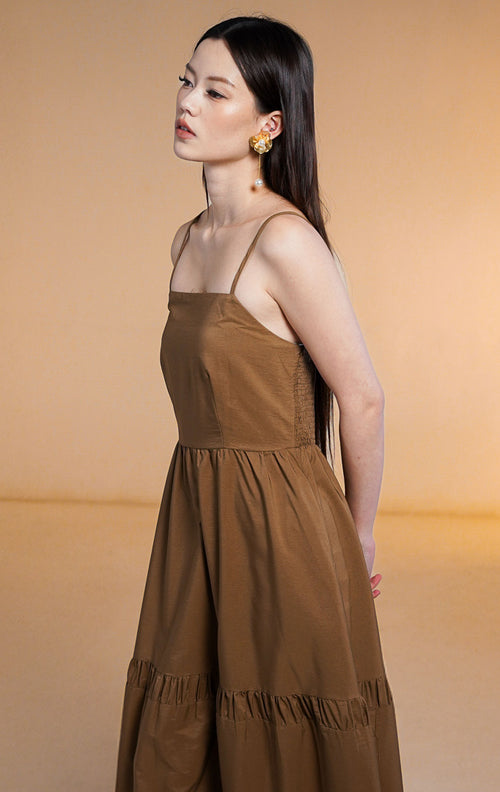 Model wanita mengenakan maxi dress susun (tiered dress) warna cokelat tua (dark brown) dengan tali spageti dan saku samping, dipadukan dengan anting bunga emas.