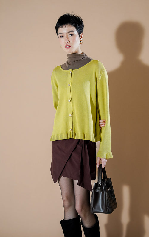 Model wanita mengenakan Angelita Cardigan berwarna Lime (hijau kekuningan) dengan detail ruffle di bagian pinggir dan kancing emas yang elegan dari This Is April.